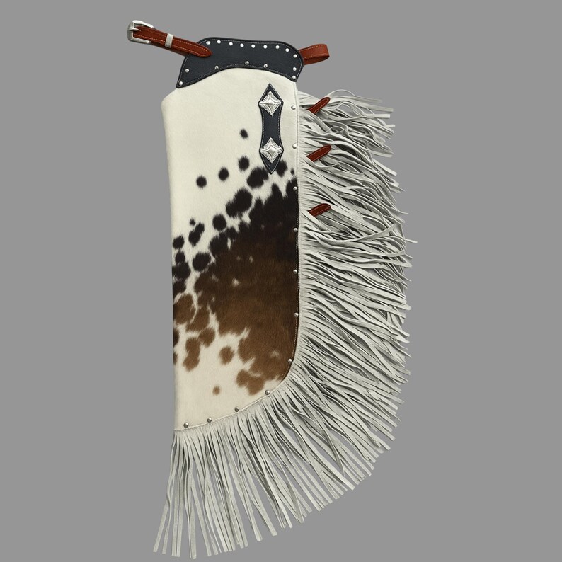 May include: Cowhide chaps with a white and brown spotted pattern. The chaps feature a black leather top with silver studs and a brown leather belt. White fringe adorns the side and bottom edges, adding a decorative element to the western-style garment.