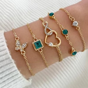Peut inclure: Cinq bracelets dorés avec différents charms : un papillon, une pierre carrée vert émeraude, un cœur et des pierres rondes vert émeraude et transparentes. Les bracelets sont fins et présentent un design en chaîne.