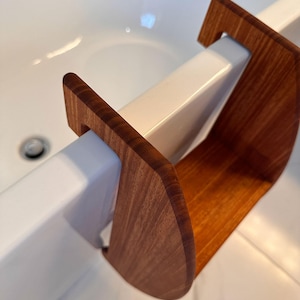May include: A wooden bath tray with a curved design, featuring a warm, reddish-brown hue. The tray is designed to rest across the edge of a white bathtub, providing a convenient surface for bath accessories. The wood grain is visible.