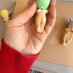 May include: A hand holding a small figurine with a green head and a nude-coloured body. The figurine has small white wings. Two other similar figurines are visible in the background, one with a yellow head and the other with a blue hat.