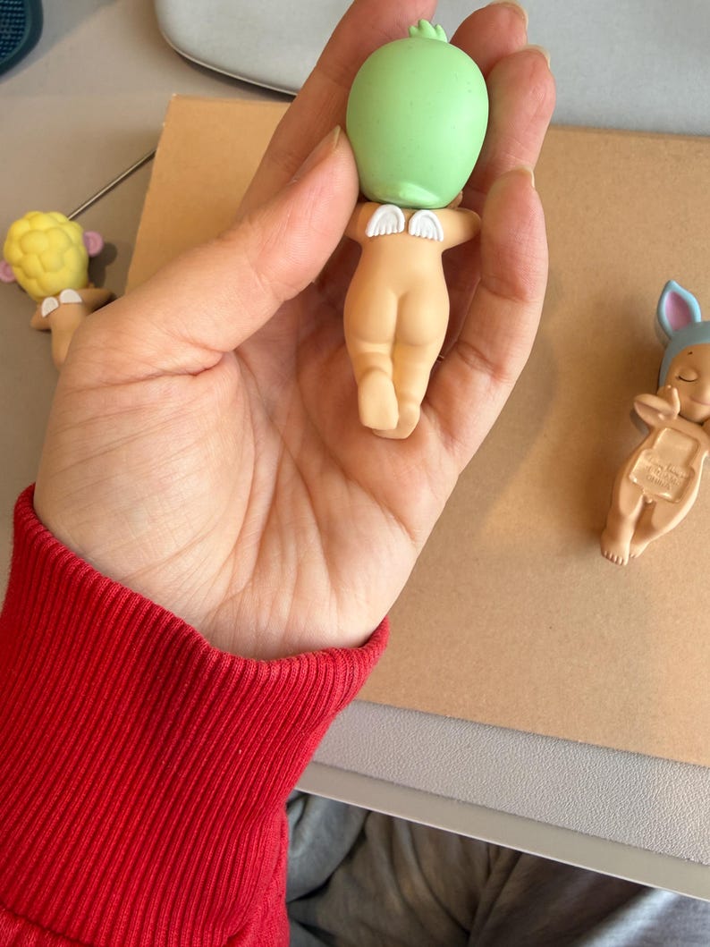 May include: A hand holding a small figurine with a green head and a nude-coloured body. The figurine has small white wings. Two other similar figurines are visible in the background, one with a yellow head and the other with a blue hat.