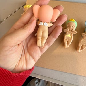 May include: A selection of small, nude-coloured figurines with angel wings and large, rounded ears. One figurine is held in a hand, while others are arranged on a light brown surface. The figurines have a minimalist design.