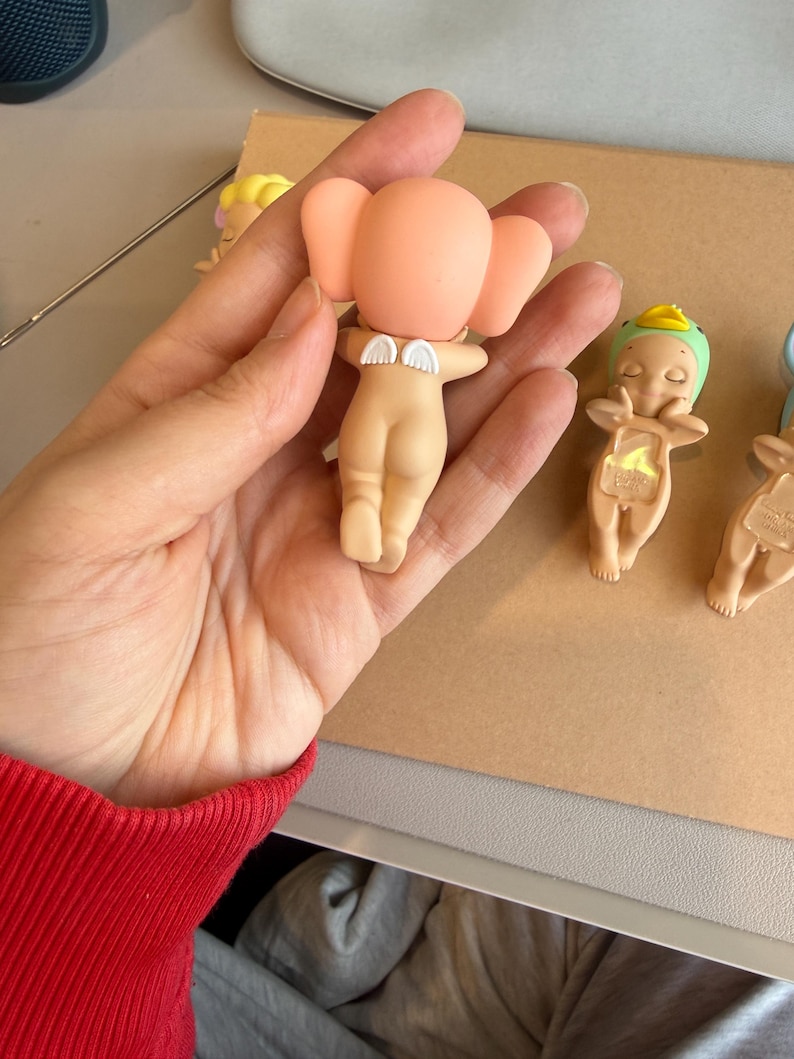 May include: A selection of small, nude-coloured figurines with angel wings and large, rounded ears. One figurine is held in a hand, while others are arranged on a light brown surface. The figurines have a minimalist design.