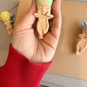 May include: A small, beige figurine of a baby wearing a green duck hat with a yellow beak. The figurine has closed eyes and is held in a hand. Other similar figurines are visible in the background.
