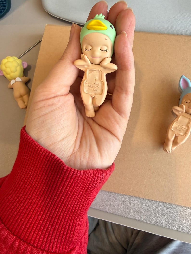 May include: A small, beige figurine of a baby wearing a green duck hat with a yellow beak. The figurine has closed eyes and is held in a hand. Other similar figurines are visible in the background.