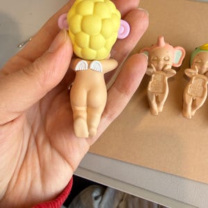 May include: A small, nude-coloured figurine with a yellow head, pink ears, and small white wings. The figurine is held in a hand, with two other similar figurines visible in the background. The figurines have a baby-like appearance.