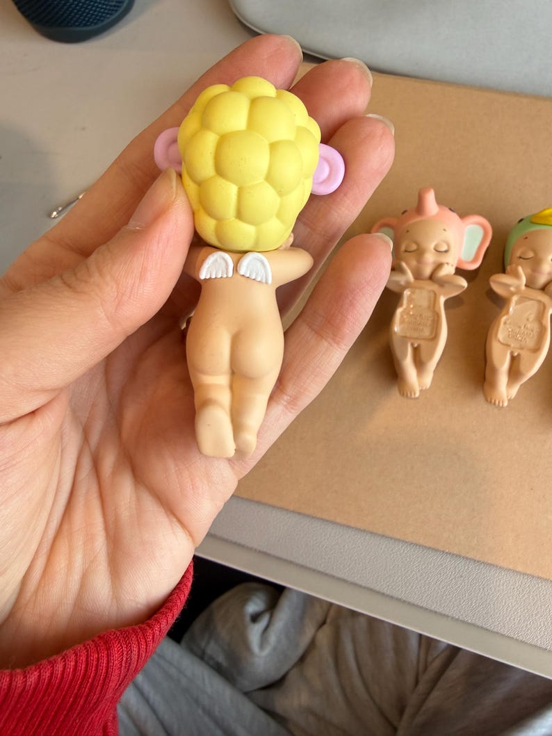 May include: A small, nude-coloured figurine with a yellow head, pink ears, and small white wings. The figurine is held in a hand, with two other similar figurines visible in the background. The figurines have a baby-like appearance.