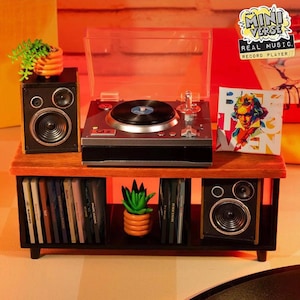 Op de afbeelding: Een miniatuur platenspelerset met een grijze draaitafel met een doorzichtig deksel, twee zwarte luidsprekers, een plank in houtstijl en een verzameling kleine vinylplaten. Een kleine potplant en een vierkante albumhoes met een portret zijn inbegrepen.