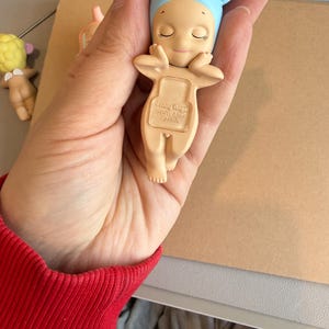 May include: A small, beige figurine of a baby wearing a blue bonnet with pink-lined ears. The baby has closed eyes and is holding its arms up. The text "Sonny Angel DREAMS CHINA" is printed on the back. Another similar figurine is visible in the background.