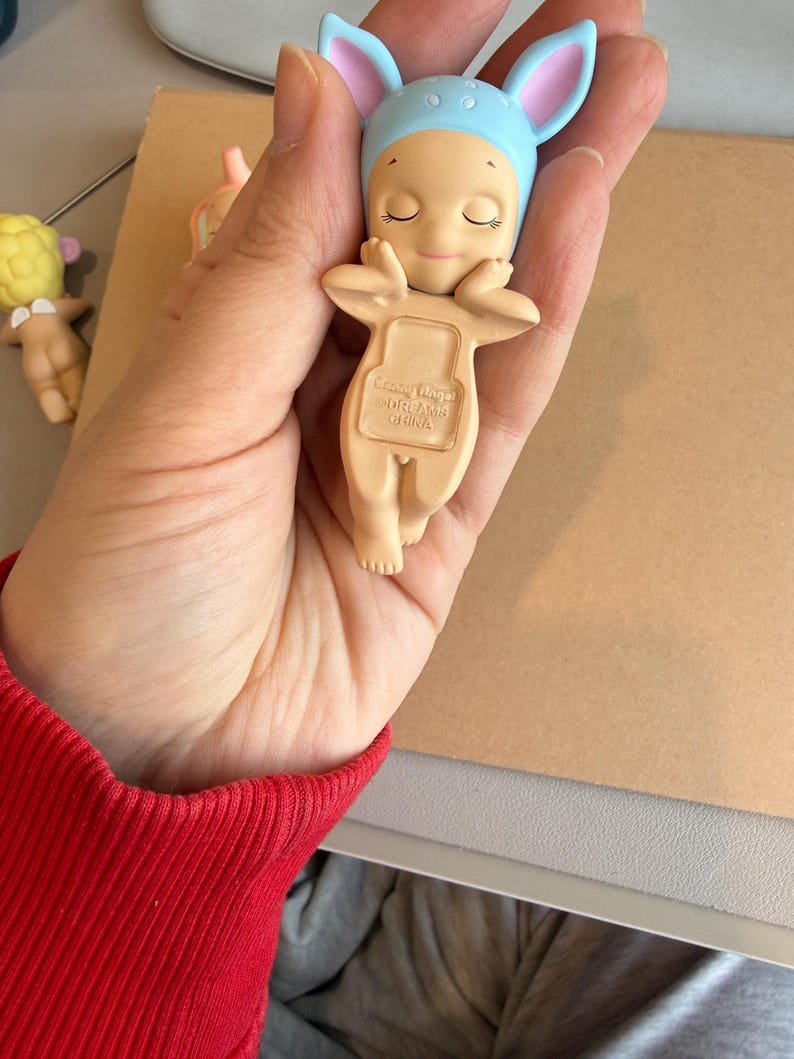 May include: A small, beige figurine of a baby wearing a blue bonnet with pink-lined ears. The baby has closed eyes and is holding its arms up. The text "Sonny Angel DREAMS CHINA" is printed on the back. Another similar figurine is visible in the background.