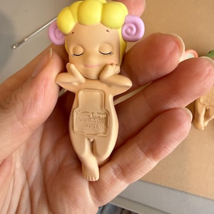 May include: A small, beige figurine of a cherubic figure with closed eyes, yellow hair, and pink accents. The figurine is holding its arms up, with the text "Sonny Angel Dreams China" visible on the back.