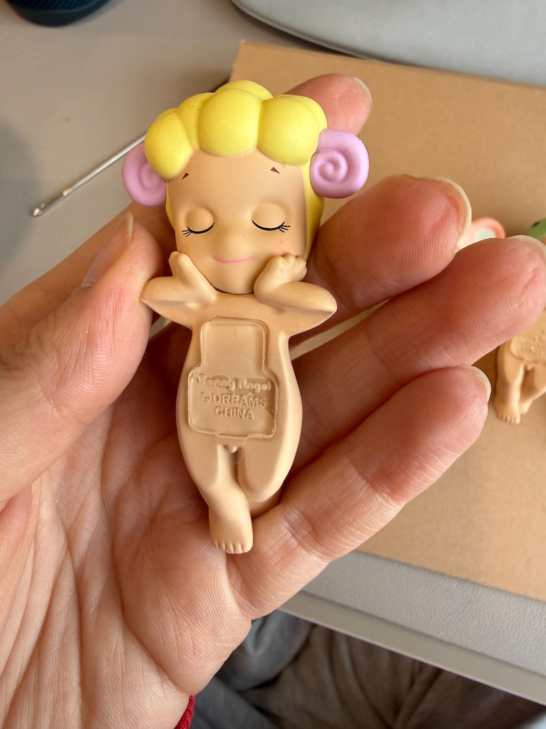 May include: A small, beige figurine of a cherubic figure with closed eyes, yellow hair, and pink accents. The figurine is holding its arms up, with the text "Sonny Angel Dreams China" visible on the back.