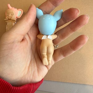 May include: A small figurine with a light blue head and large ears, small white wings, and a nude-coloured body. The figurine is held in a hand. Another figurine with a pink elephant head is visible in the background.