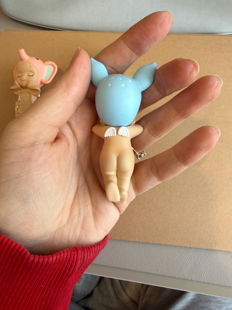 May include: A small figurine with a light blue head and large ears, small white wings, and a nude-coloured body. The figurine is held in a hand. Another figurine with a pink elephant head is visible in the background.