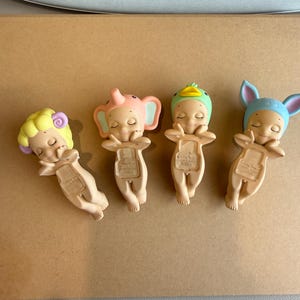 May include: Four small figurines of sleeping babies, each with an animal-themed hat: a sheep, an elephant, a duck, and a bunny. The figurines are beige with pastel colours.