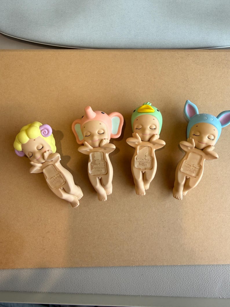 May include: Four small figurines of sleeping babies, each with an animal-themed hat: a sheep, an elephant, a duck, and a bunny. The figurines are beige with pastel colours.