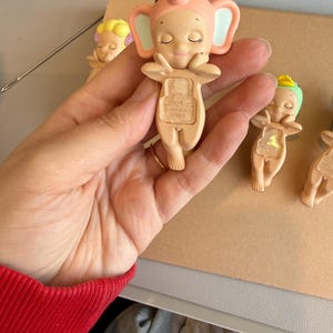 May include: Several small, light-tan figurines are visible. The main figurine is an elephant with a pink hat and ears, arms crossed, and closed eyes. The base of the figurine has the text "DREAMS CHINA". Other figurines are in the background.