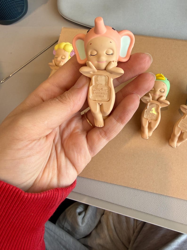 May include: Several small, light-tan figurines are visible. The main figurine is an elephant with a pink hat and ears, arms crossed, and closed eyes. The base of the figurine has the text "DREAMS CHINA". Other figurines are in the background.