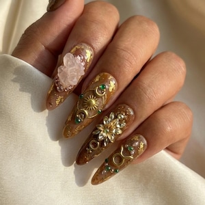 May include: Long, almond-shaped nails with a brown base, adorned with gold leaf, crystals, and intricate gold embellishments. The designs include a sun, moon, and star motif, along with floral and zodiac symbols. The nails are a unique fashion statement.