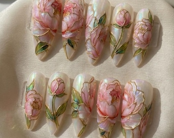 May include: A set of ten long, almond-shaped artificial nails. Each nail is painted with a pearlescent base and features a detailed pink peony design with green leaves and gold outlines. The floral design is hand-painted.