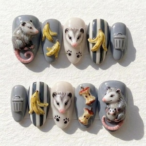 May include: A set of ten artificial nails in grey, white, and yellow. The nails feature 3D possum, banana, apple core, and bin designs. The possums are grey and white with pink noses and tails. The bananas are yellow.