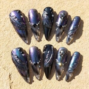 May include: A set of ten long, pointed false nails. The nails feature a dark iridescent color scheme with purple, blue, and black hues. Some nails have feather designs, while others have rhinestones or a skull design.