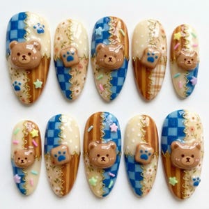 May include: A set of ten press-on nails featuring a split design with blue checkered patterns, floral prints, and brown stripes. Each nail is decorated with a 3D brown bear face, small star-shaped charms, and tiny blue paw print accents.