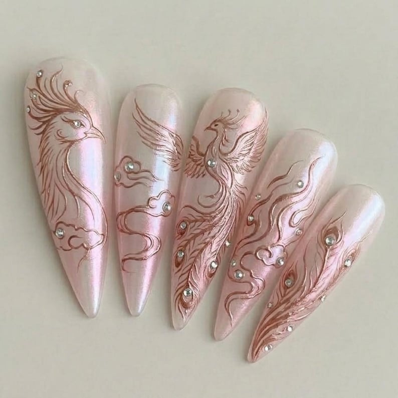May include: Five long, almond-shaped artificial nails with a pearlescent pink finish. Each nail features a copper-colored phoenix design, some with cloud accents and small crystal embellishments. The phoenix designs vary across the nails.