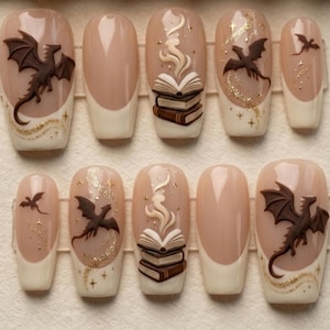May include: A set of ten press-on nails featuring a nude base with cream-colored French tips. Designs include raised 3D brown dragons, stacks of books with swirling smoke, and gold glitter accents in the shape of stars and flowing trails.