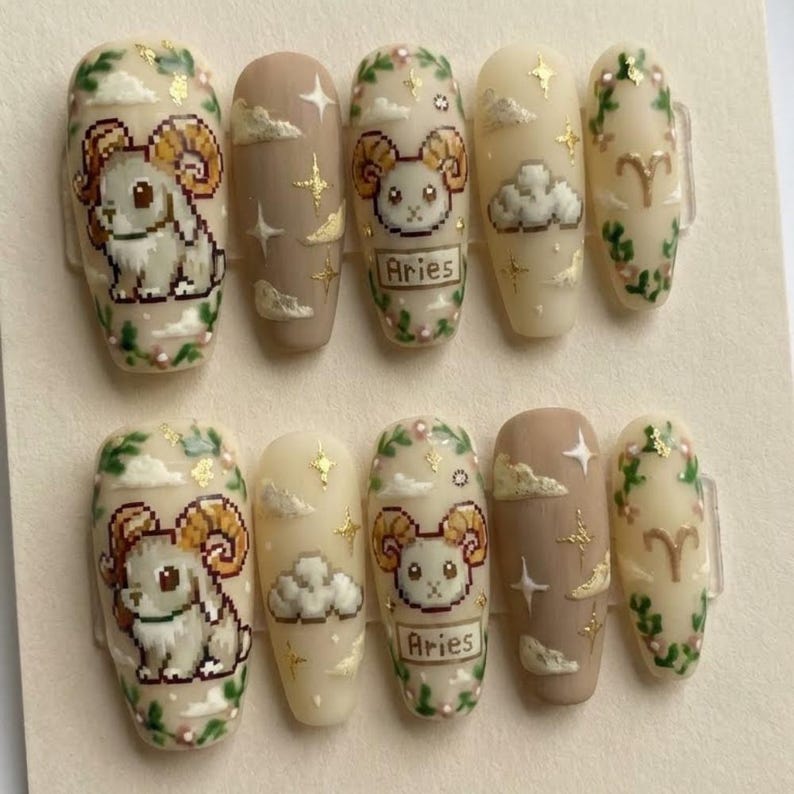 Puede incluir: Un conjunto de diez u&ntilde;as postizas con tem&aacute;tica de Aries. Las u&ntilde;as presentan ilustraciones pixeladas de carneros, nubes, estrellas y detalles florales en beige, marr&oacute;n y dorado. La palabra "Aries" est&aacute; impresa en algunas u&ntilde;as.
