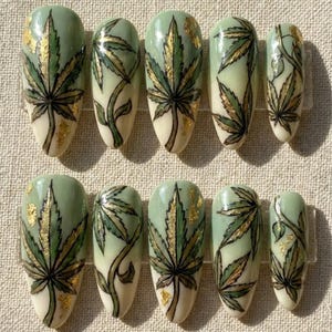 Puede incluir: Un conjunto de diez uñas postizas en forma de almendra. Cada uña está pintada con una base verde claro, una punta blanca y un diseño detallado de hoja de cannabis en verde y dorado. Las hojas tienen contornos negros.