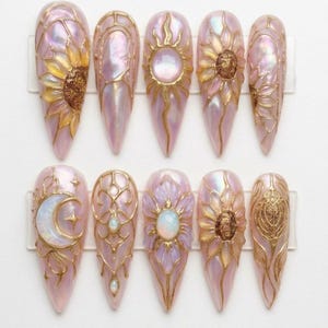 May include: A set of ten long, almond-shaped artificial nails. Each nail features a pearlescent pink base with gold-colored embellishments. Designs include sunflowers, sunbursts, moons, and floral patterns, creating a unique and artistic aesthetic.