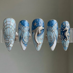K&ouml;nnte beinhalten: F&uuml;nf blaue und silberne Nageldesigns. Eines zeigt eine Muschel, zwei haben Wellenmuster, eines einen Delfin und das letzte ein Seepferdchen. Jeder Nagel hat einen silbernen Metallakzent und ein gl&auml;nzendes Finish.
