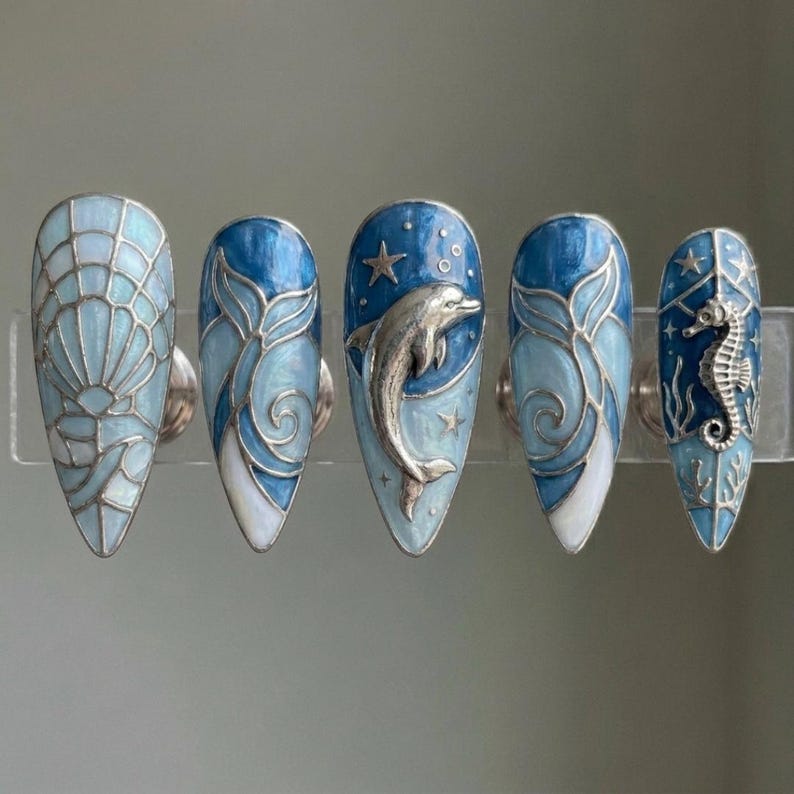 K&ouml;nnte beinhalten: F&uuml;nf blaue und silberne Nageldesigns. Eines zeigt eine Muschel, zwei haben Wellenmuster, eines einen Delfin und das letzte ein Seepferdchen. Jeder Nagel hat einen silbernen Metallakzent und ein gl&auml;nzendes Finish.