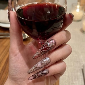 Peut inclure: Une main tenant un verre de vin rouge, avec de longs ongles décorés. Les ongles sont ornés de motifs argentés, rouges et roses. Le verre à vin est transparent et le vin est d'une couleur rouge foncé. Les ongles ont un design élaboré et orné.