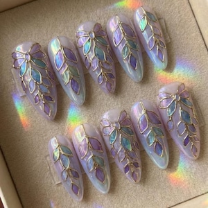 May include: A set of ten artificial nails with a pearlescent finish. Each nail features a gold-colored bow and leaf design with iridescent purple, blue, and green accents. The nails are almond-shaped and arranged in a display.