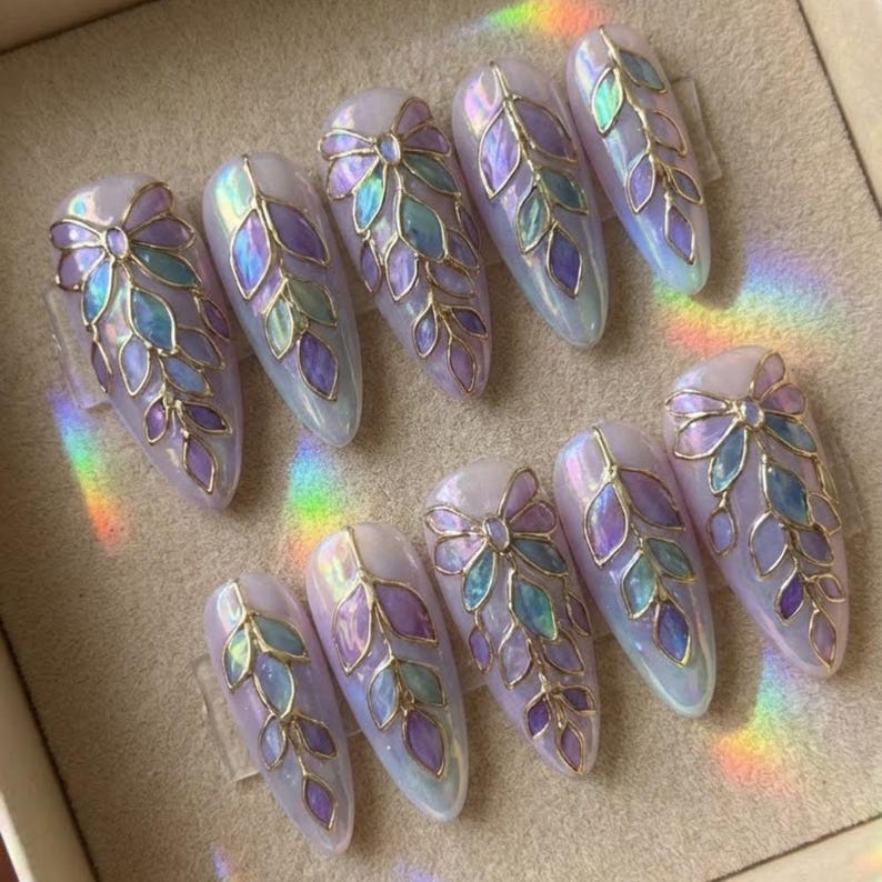 May include: A set of ten artificial nails with a pearlescent finish. Each nail features a gold-colored bow and leaf design with iridescent purple, blue, and green accents. The nails are almond-shaped and arranged in a display.