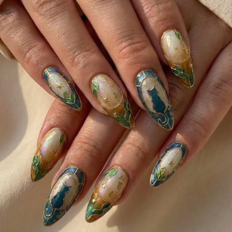 May include: Almond-shaped nail art with a gold trim and intricate designs. The nails feature a cat silhouette, gold stars, and green leaf accents. The color palette includes teal, gold, and iridescent white.
