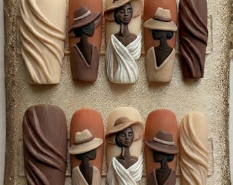 May include: A set of ten artificial nails with a neutral color palette. The nails feature 3D art of women wearing hats and draped fabric. Some nails have a textured, swirled design in shades of brown and beige.