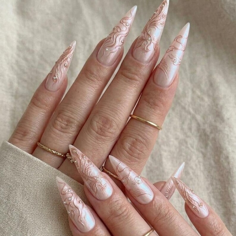 May include: Long, pointed, artificial nails with a pearlescent finish. The nails feature intricate, swirling designs in a rose gold color, accented with small, sparkling embellishments. Gold rings adorn the fingers.