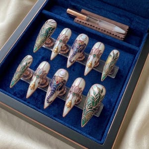 May include: A set of ten long, almond-shaped press-on nails with intricate designs. The nails feature white, gold, green, and blue patterns, including elephant and lotus motifs. The set is displayed in a blue velvet box with a small applicator.