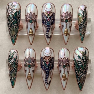 May include: A set of ten long, pointed false nails. The nails feature intricate designs, including an elephant, lotus flowers, and leaf patterns, all in shades of blue, green, and gold. Each nail has a pearlescent white base and is accented with copper-colored metal.