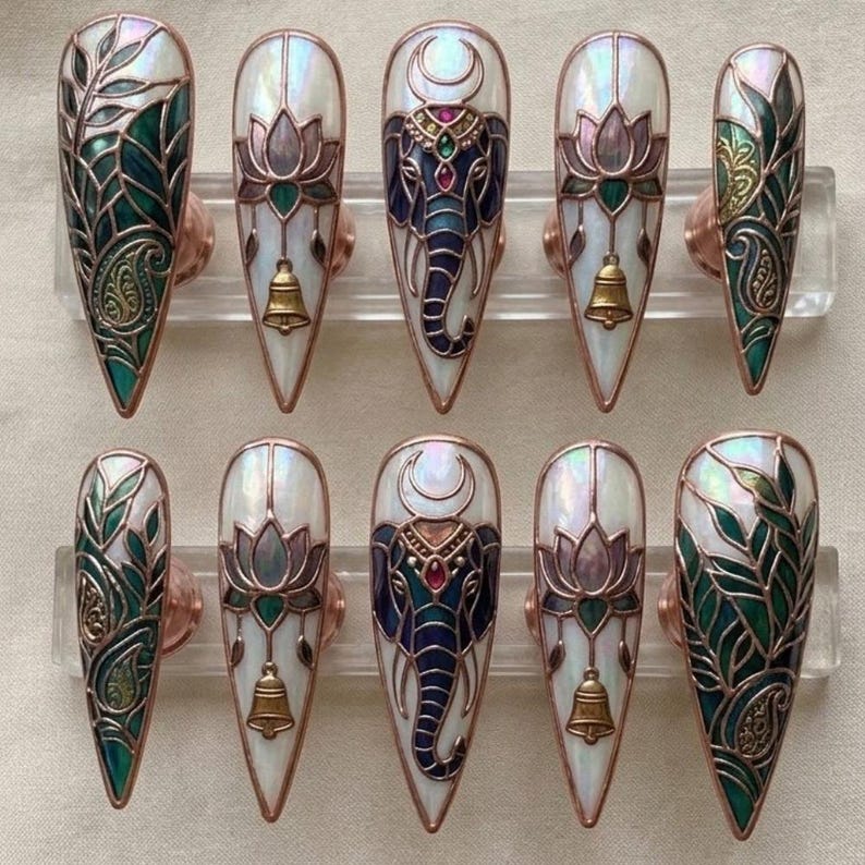 May include: A set of ten long, pointed false nails. The nails feature intricate designs, including an elephant, lotus flowers, and leaf patterns, all in shades of blue, green, and gold. Each nail has a pearlescent white base and is accented with copper-colored metal.