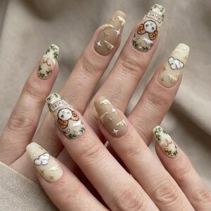 Puede incluir: Un conjunto de u&ntilde;as acr&iacute;licas largas en forma de ata&uacute;d. Las u&ntilde;as presentan una combinaci&oacute;n de colores beige y beige claro con varios dise&ntilde;os, incluyendo acentos de estrellas y nubes, y una ilustraci&oacute;n de un carnero de dibujos animados con la palabra "Aries".