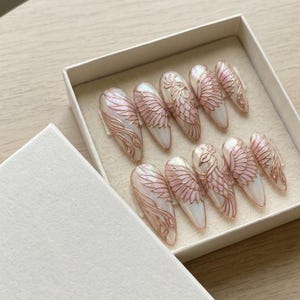 May include: A set of ten almond-shaped press-on nails in a white box. Each nail features a pearlescent white base with raised, metallic rose gold line art depicting intricate bird wings and feathers, with one nail showcasing a full bird design.