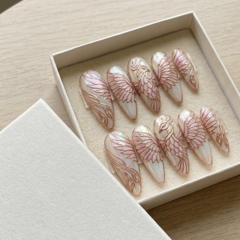 May include: A set of ten almond-shaped press-on nails in a white box. Each nail features a pearlescent white base with raised, metallic rose gold line art depicting intricate bird wings and feathers, with one nail showcasing a full bird design.