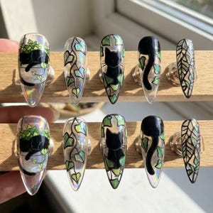 May include: A set of ten clear, long, almond-shaped press-on nails. Each nail features a unique design, including black cats, ivy, and potted plants. The designs are accented with green and silver details.