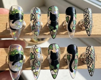 May include: A set of ten clear, long, almond-shaped press-on nails. Each nail features a unique design, including black cats, ivy, and potted plants. The designs are accented with green and silver details.