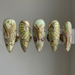 May include: Five almond-shaped, press-on nails with a light green iridescent base and gold accents. Each nail features a different design, including a feather, butterfly, and wing patterns. The nails are displayed on a clear stand.