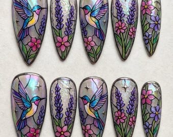 May include: Set of ten iridescent press-on nails with a stained-glass design. Each nail features a hummingbird, lavender, and pink flowers. The nails have a glossy finish and a silver outline.